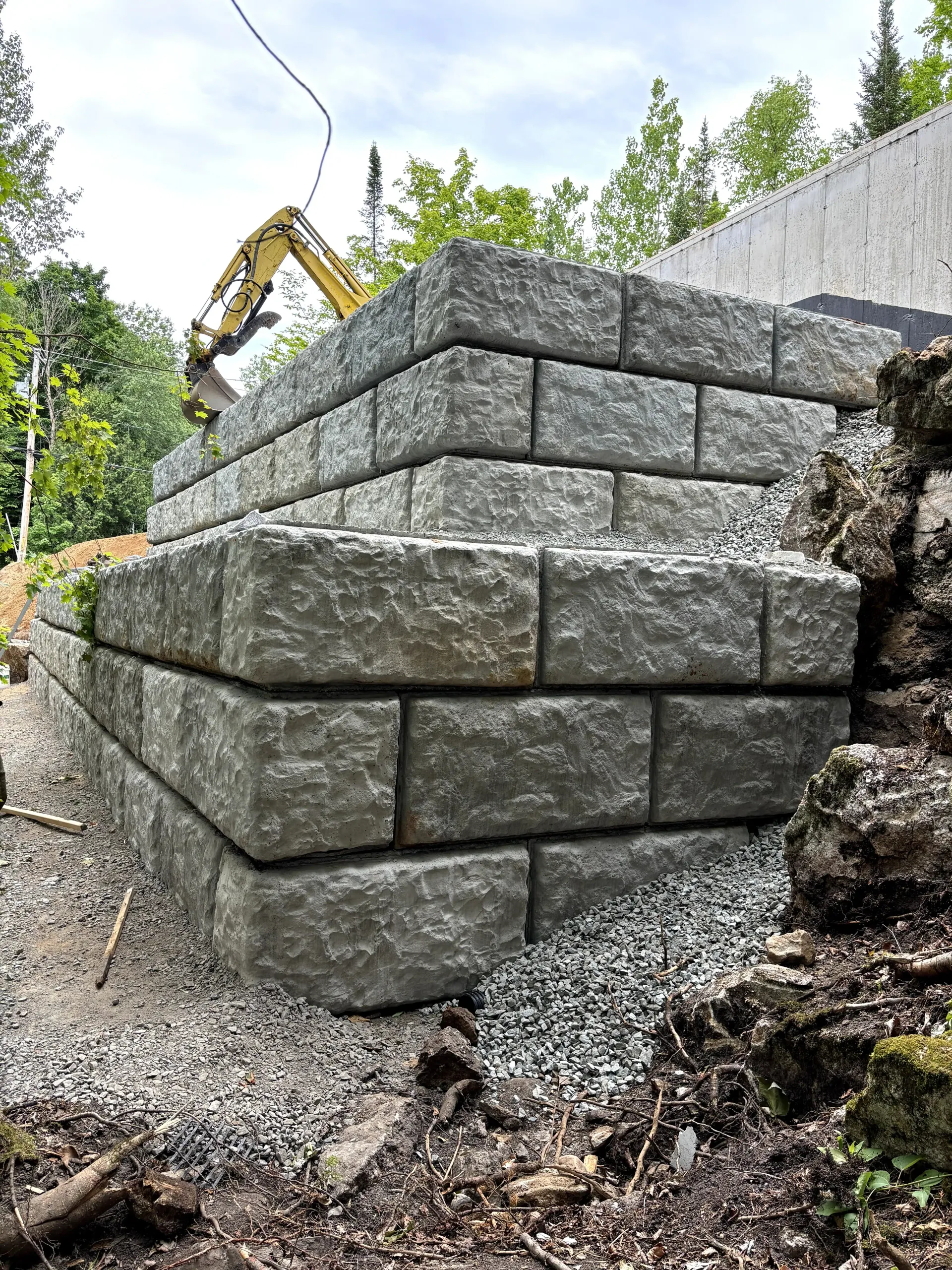 Retaining wall built from gray stone blocks beside a hillside, with an excavator in the background