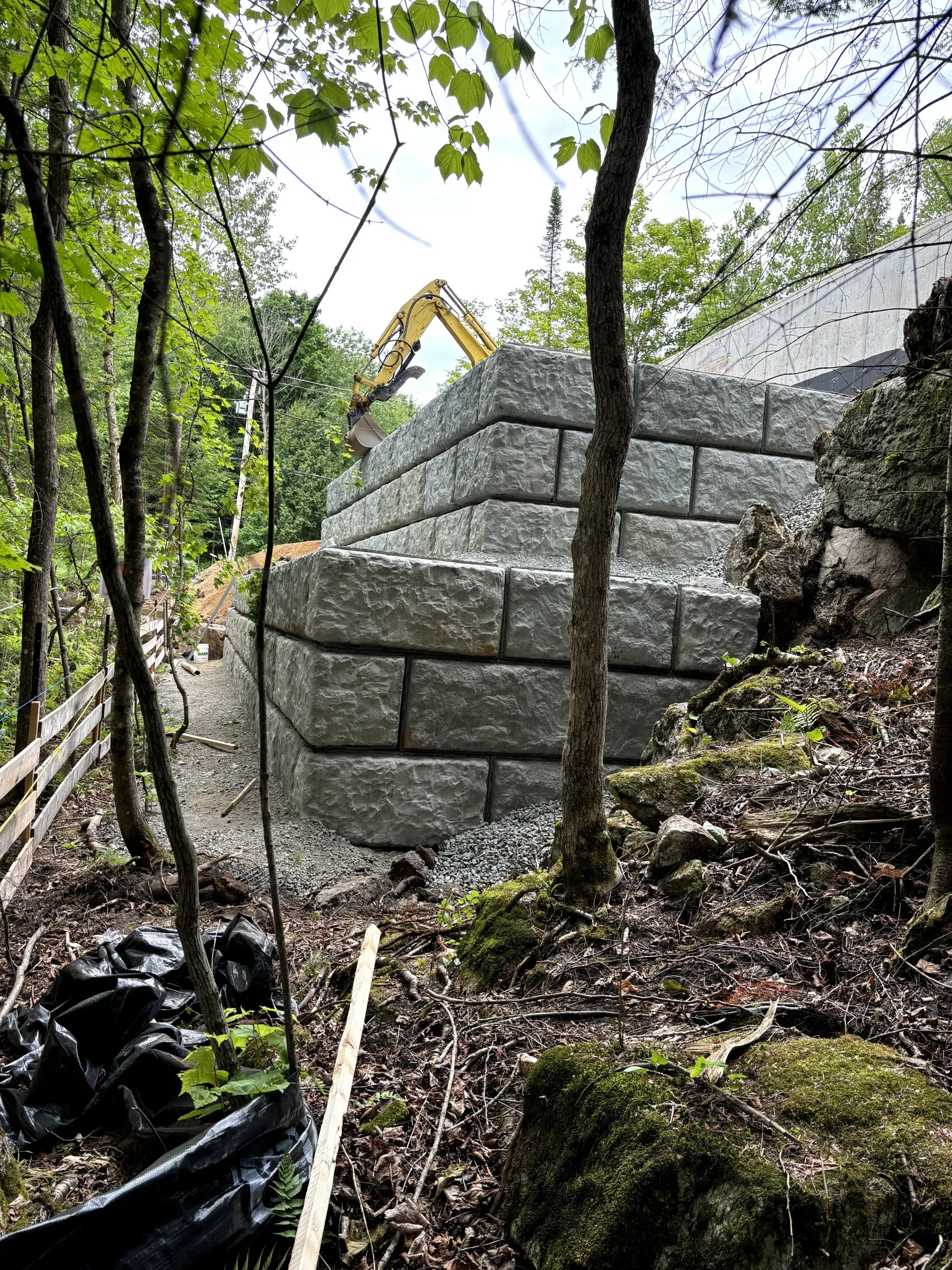 Stone retaining wall under construction on a wooded hillside, with trees and gravel piles nearby