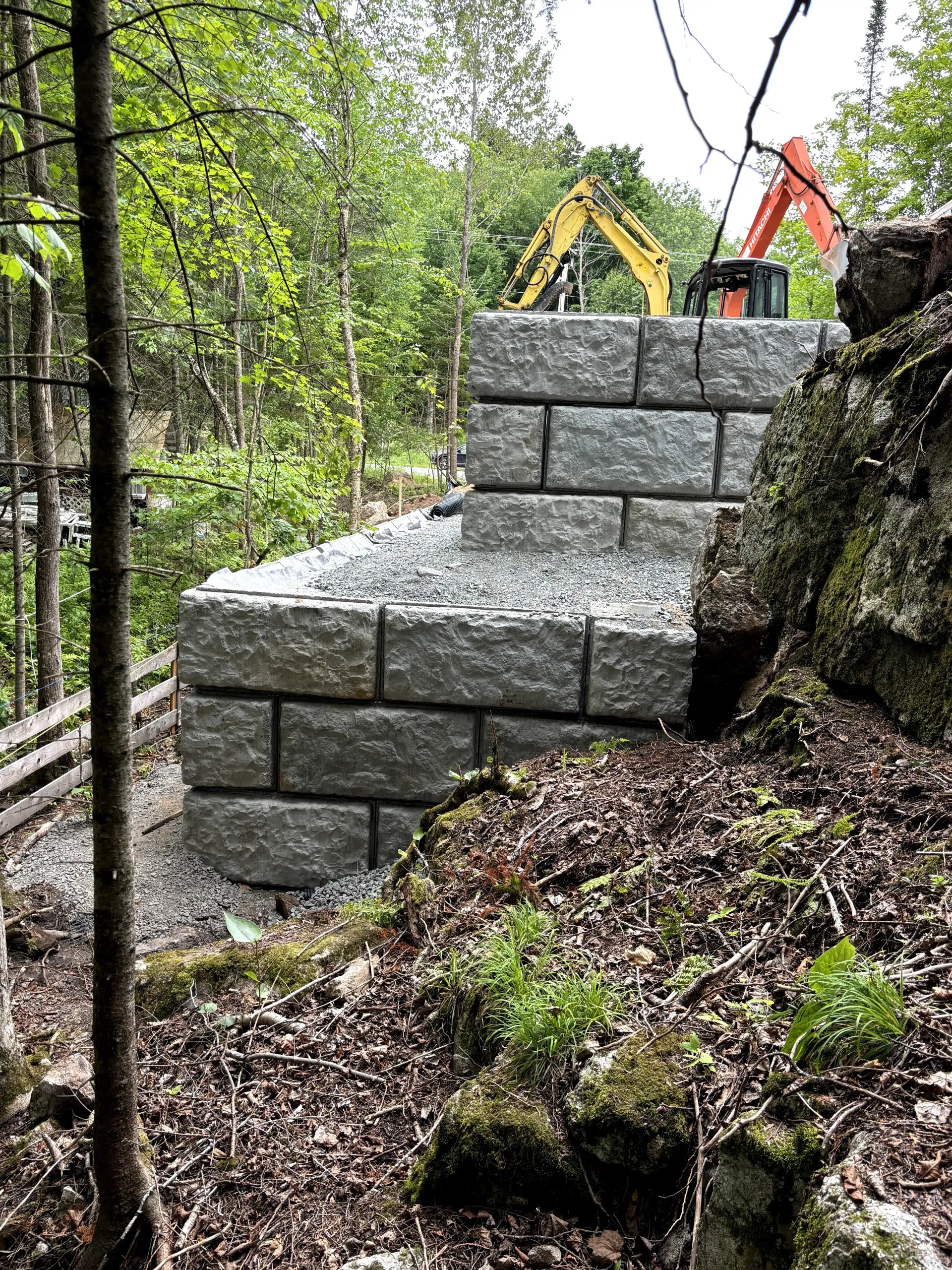 Stone retaining wall under construction on a wooded hillside, with excavators in the background.