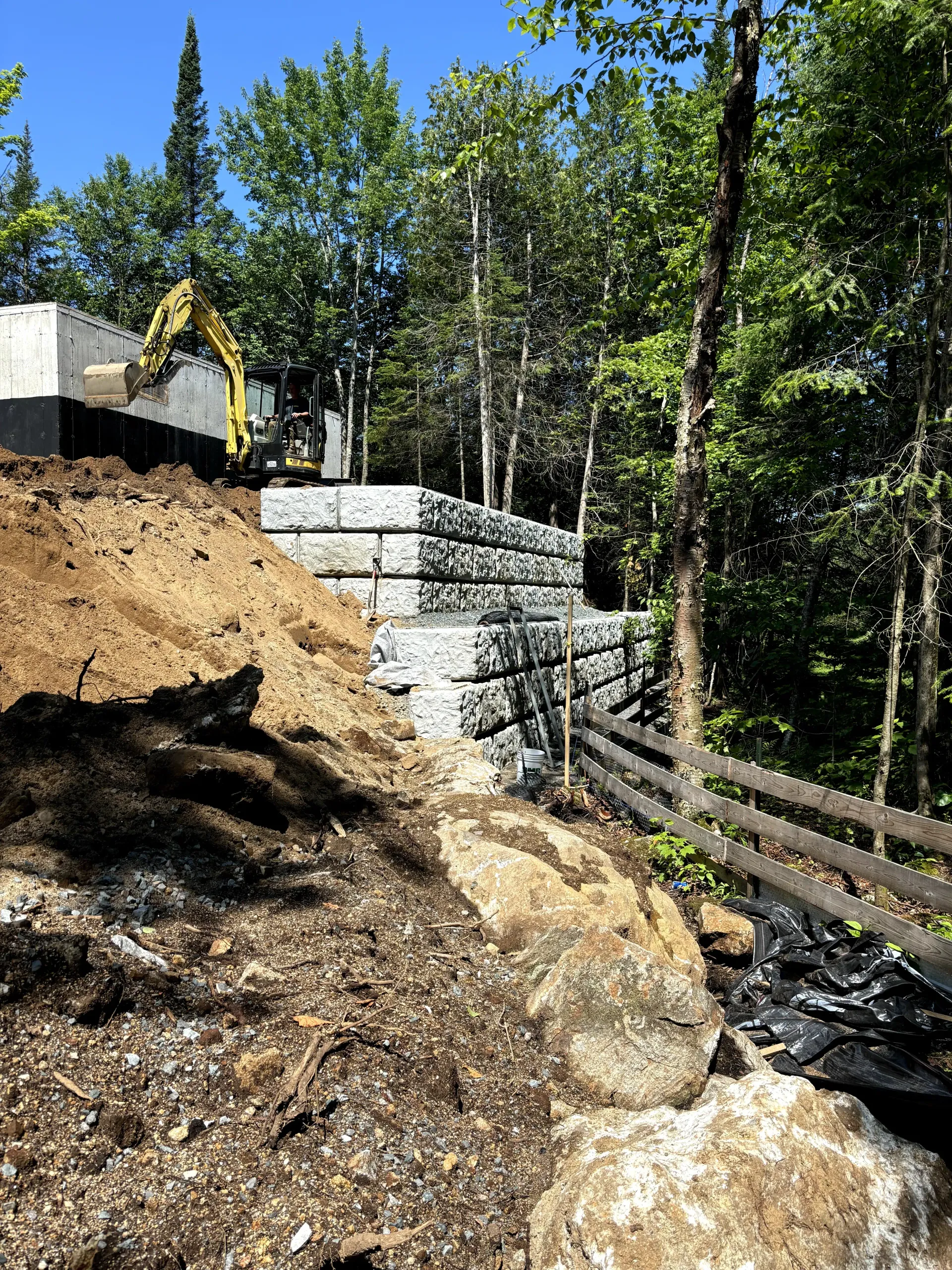 Steep dirt slope beside stacked concrete retaining wall and wooden fence in a wooded area