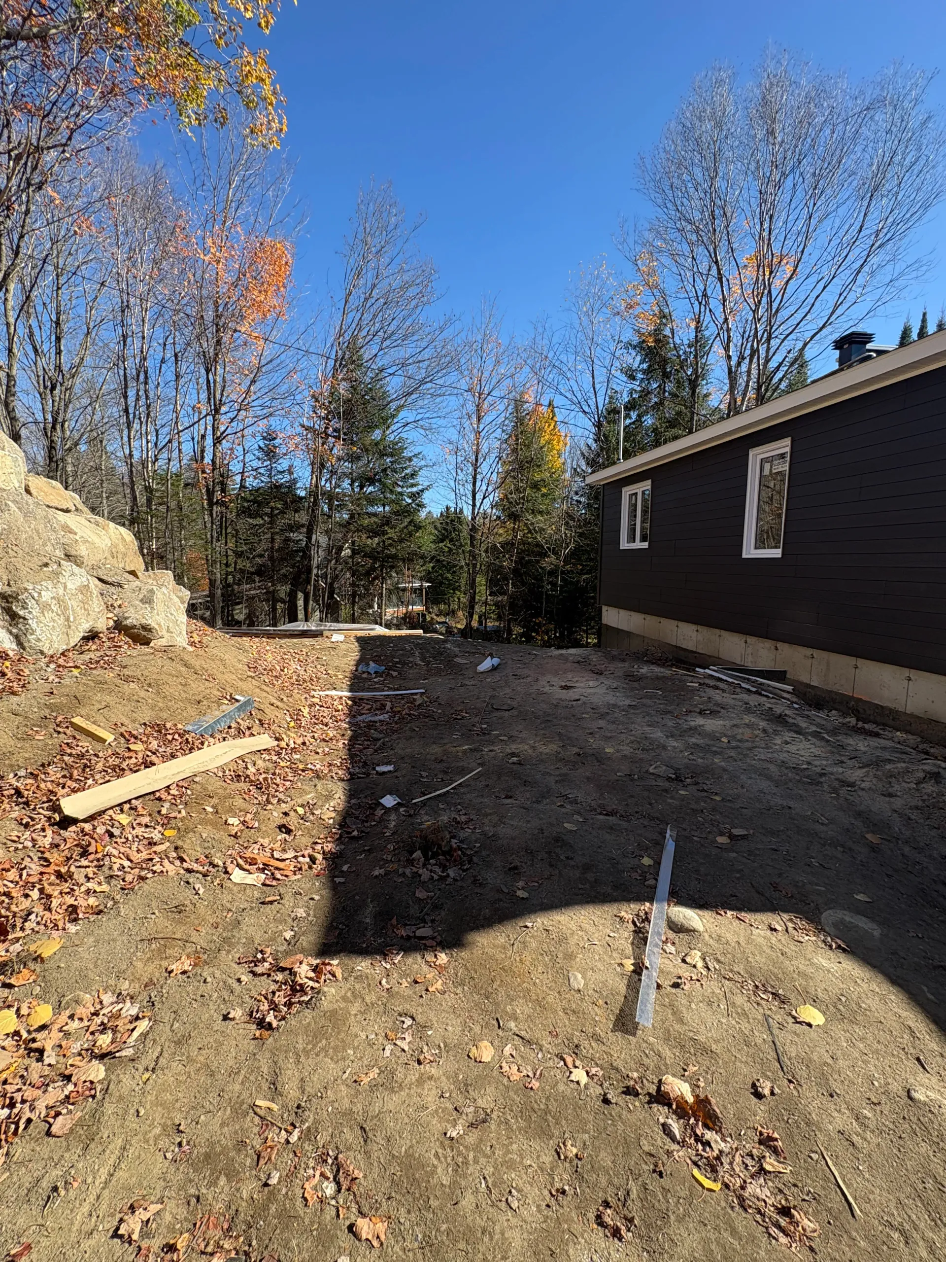 Dirt yard beside a dark house, with trees, fallen leaves, and a trench in the foreground under a blue sky.