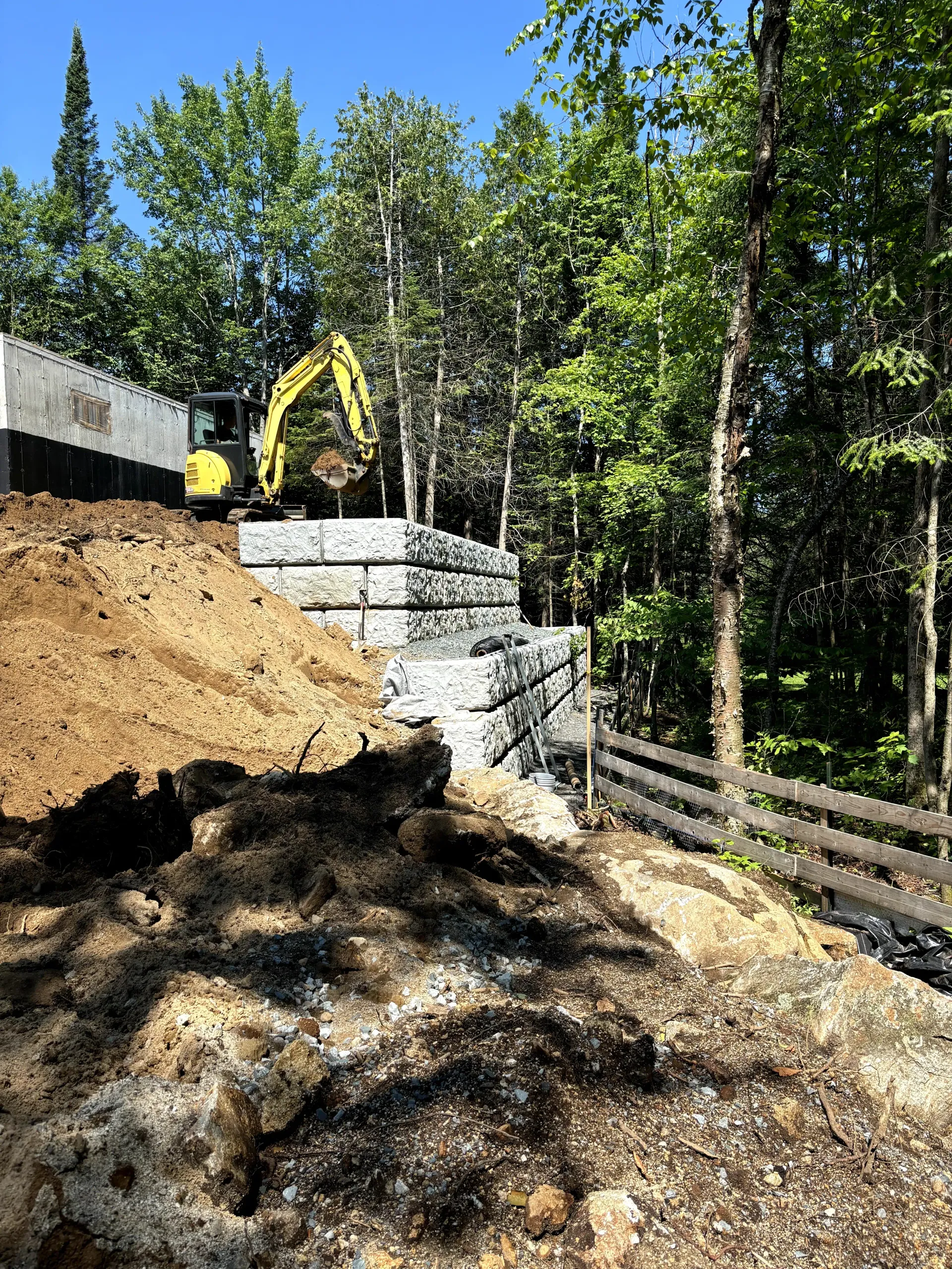 Excavator building a stone retaining wall on a wooded hillside beside a fence