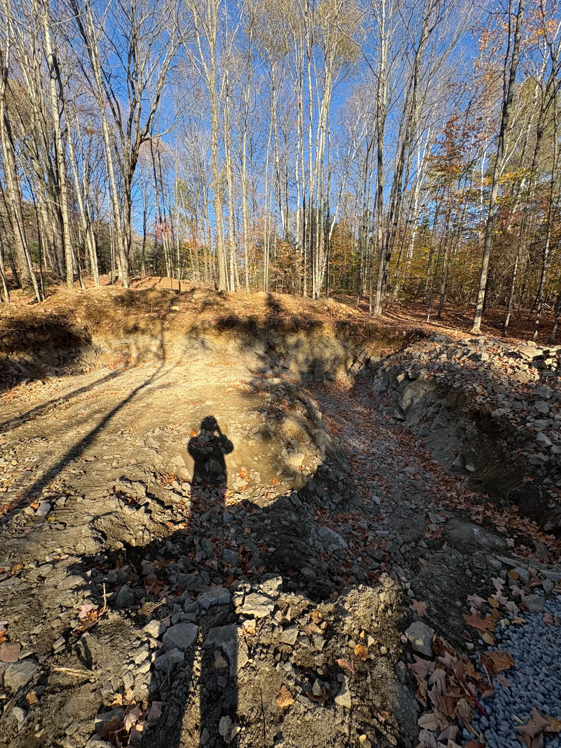 Construction site with gravel pad, white drainage pipes, and an excavator near a wooded area