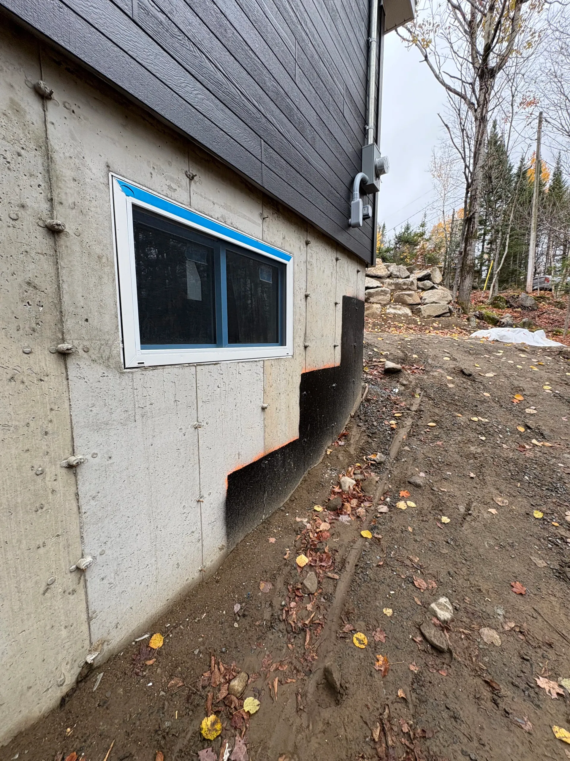 House foundation wall with a basement window and muddy ground beside a wooded slope