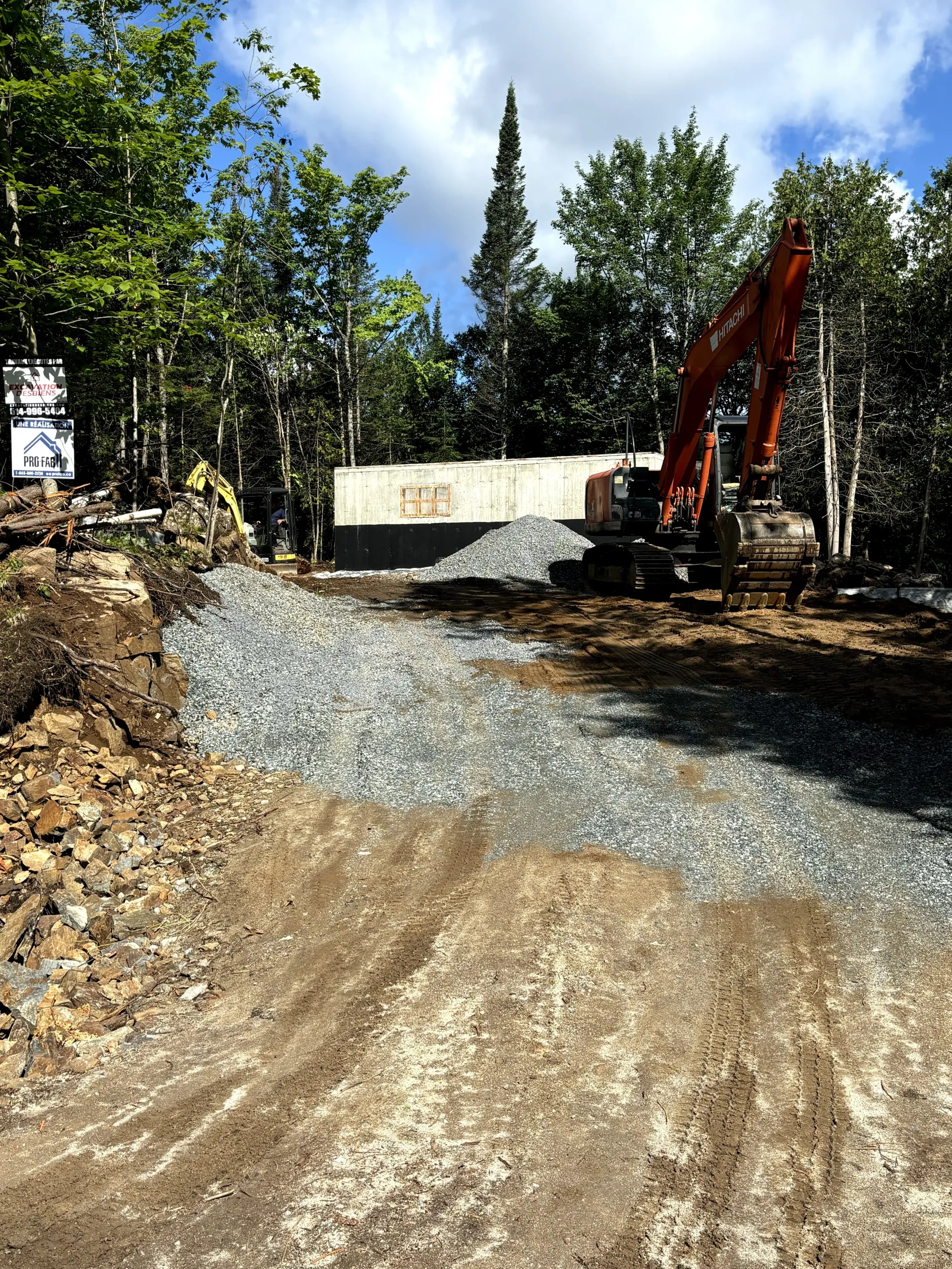 Construction site with gravel road, dirt piles, trees, and an excavator beside a concrete wall.