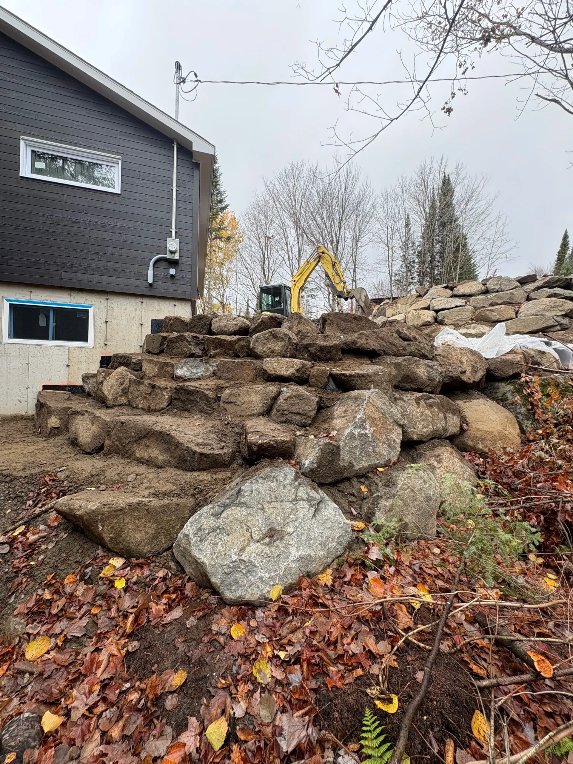 Stone retaining wall under construction beside a gray house, with an excavator in the background.