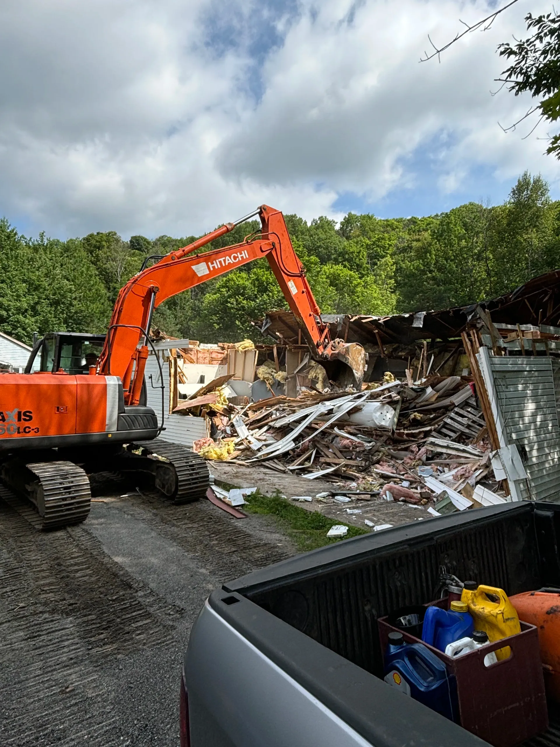 Orange excavator demolishing a collapsed house amid debris under a cloudy sky