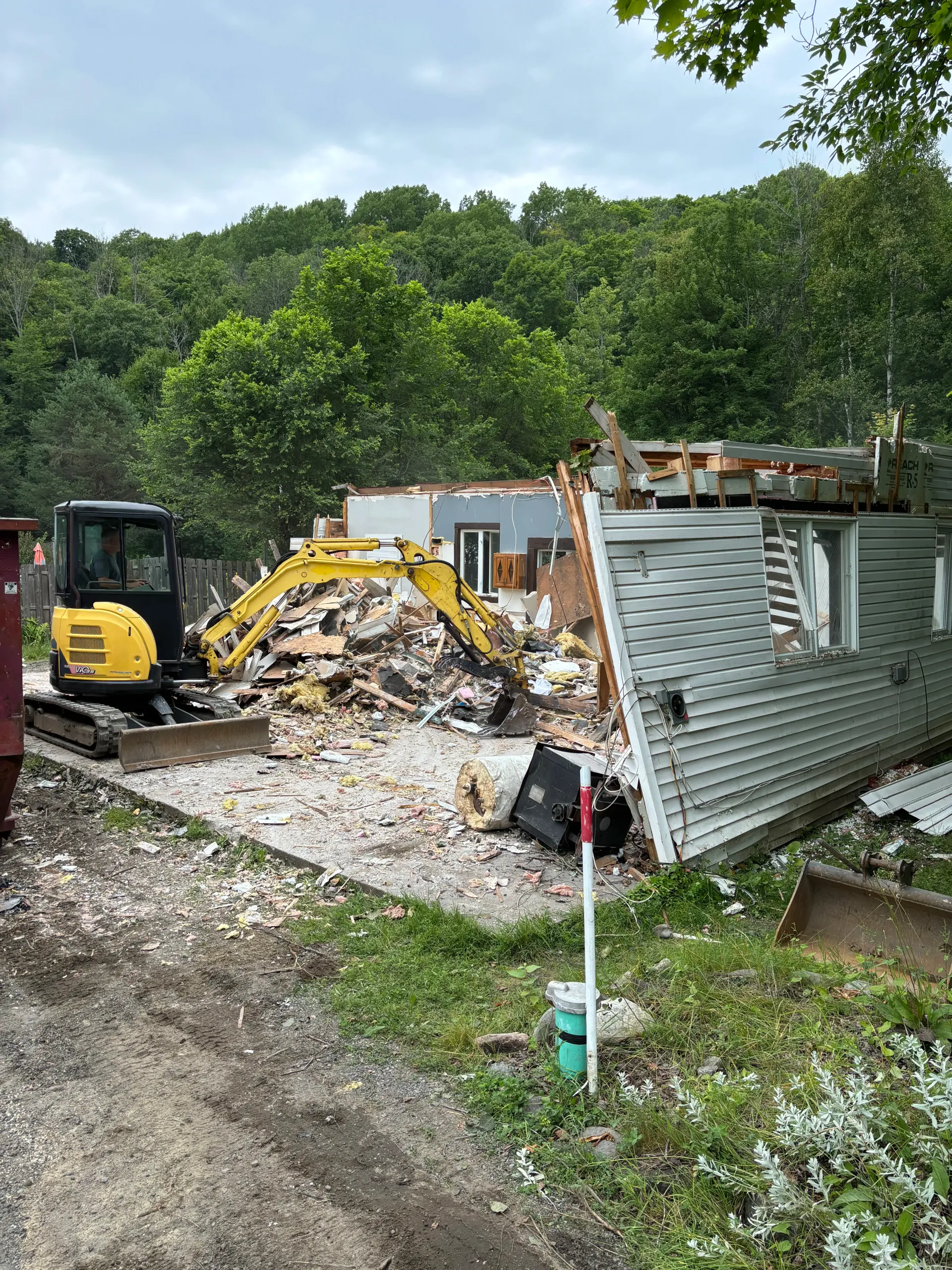 Demolition site with excavator, rubble, and a collapsed corrugated metal shed in a wooded area