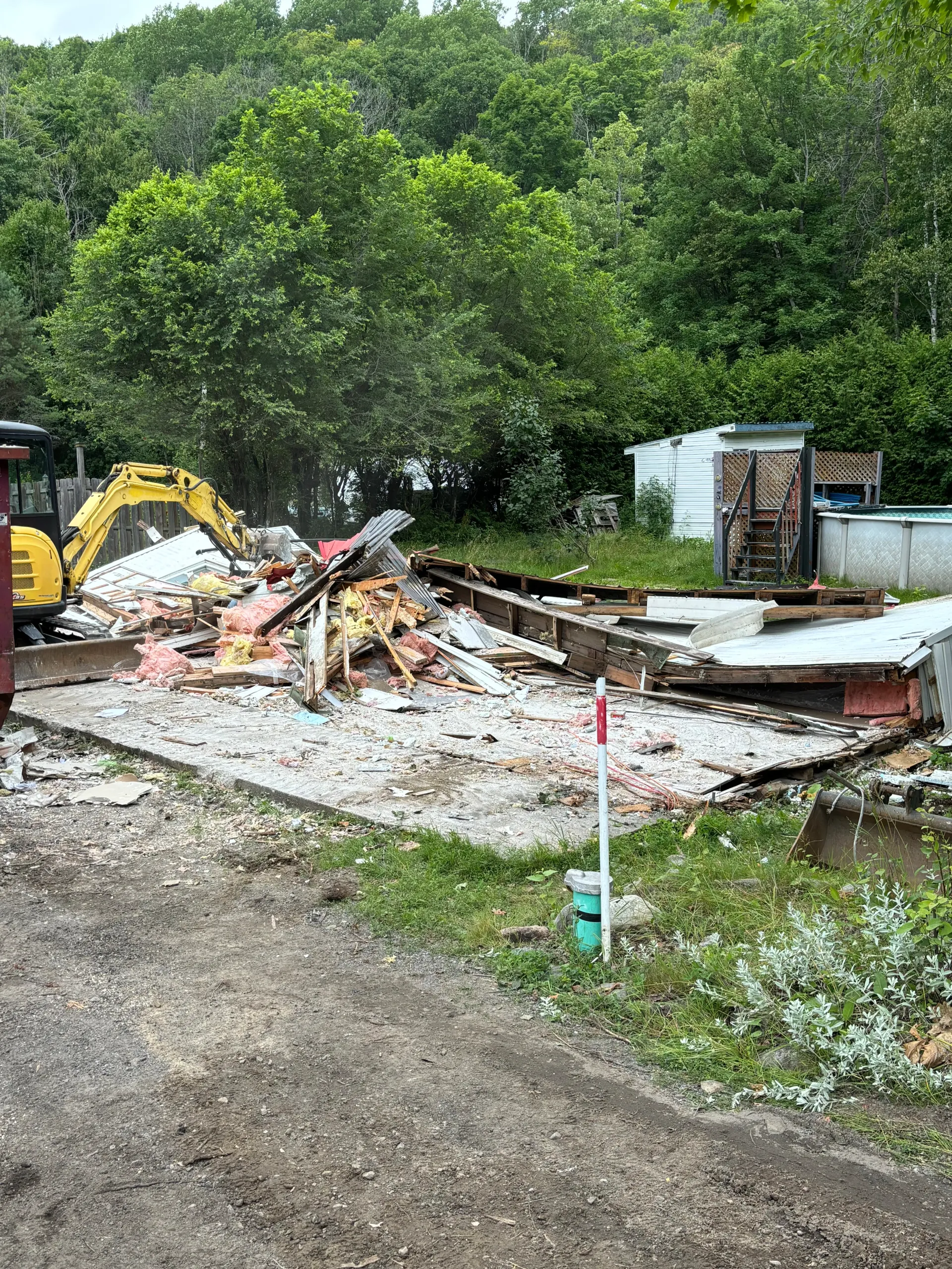 Demolition debris beside an excavator on a muddy construction site with trees and a shed in the background