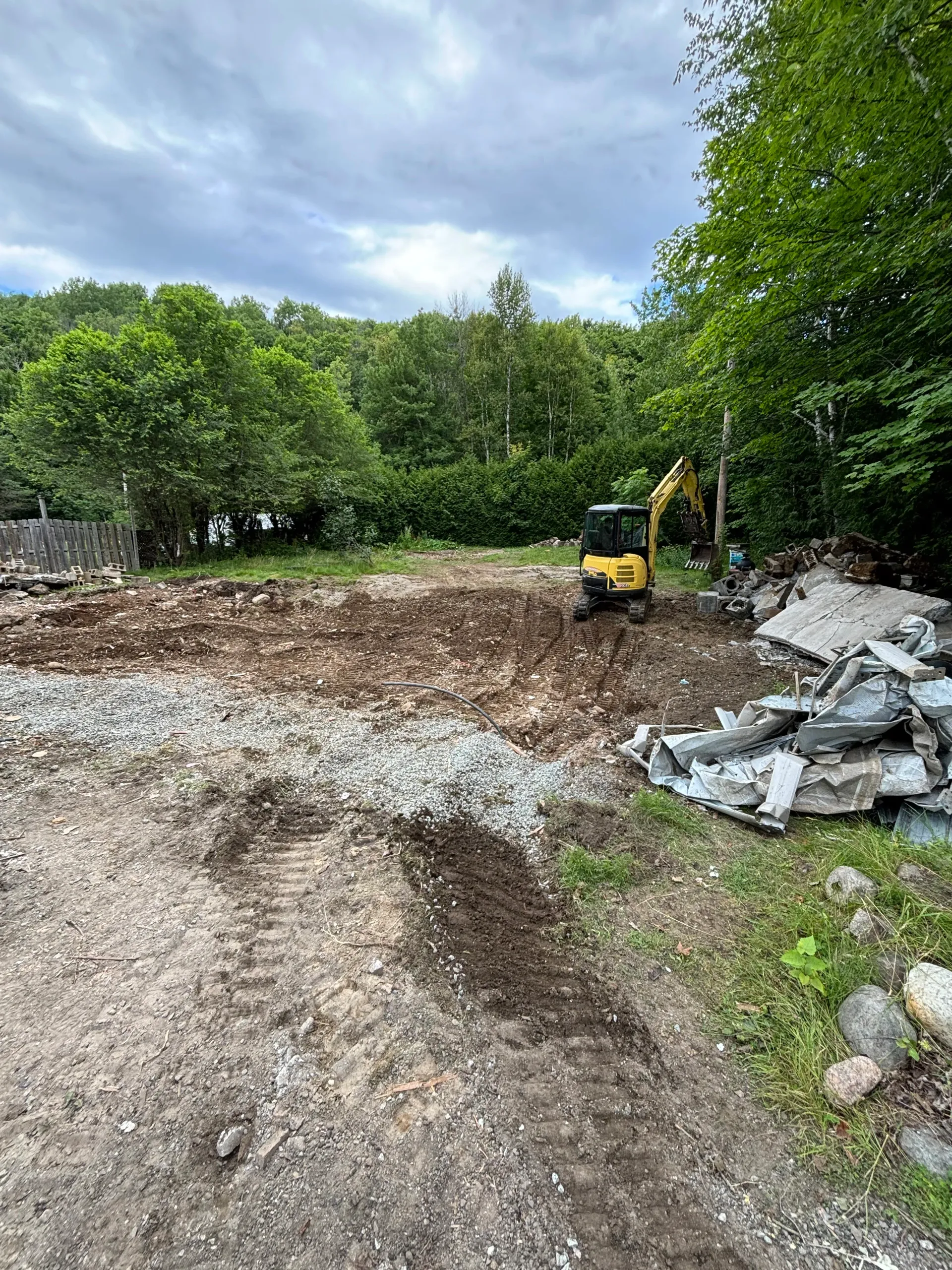 Cleared dirt lot with a small excavator, piles of rocks, and trees under a cloudy sky