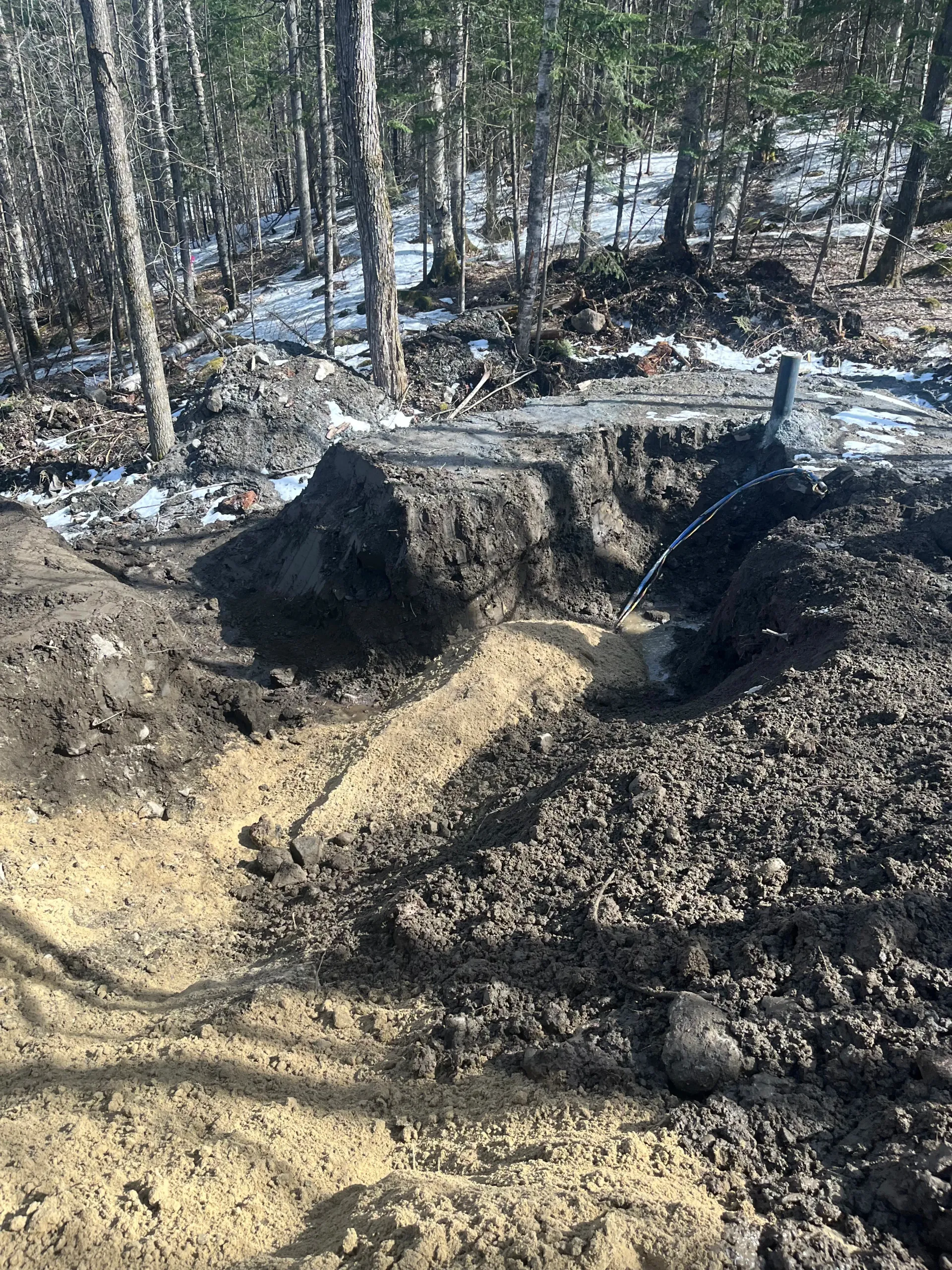 Dirt pit in a wooded area with exposed soil, snow patches, and tire tracks.