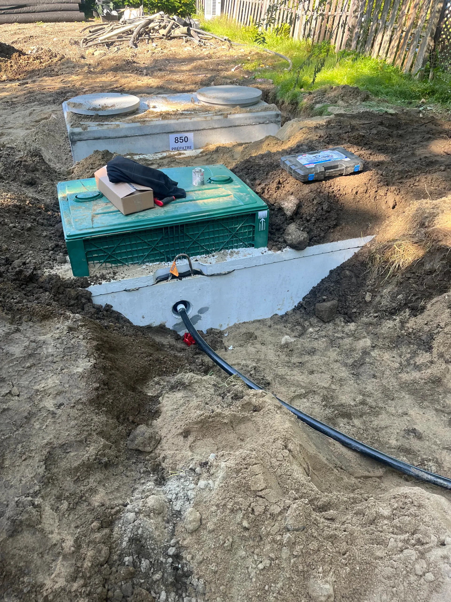 Backyard excavation with green utility box, exposed pipes, and a black hose on dirt ground
