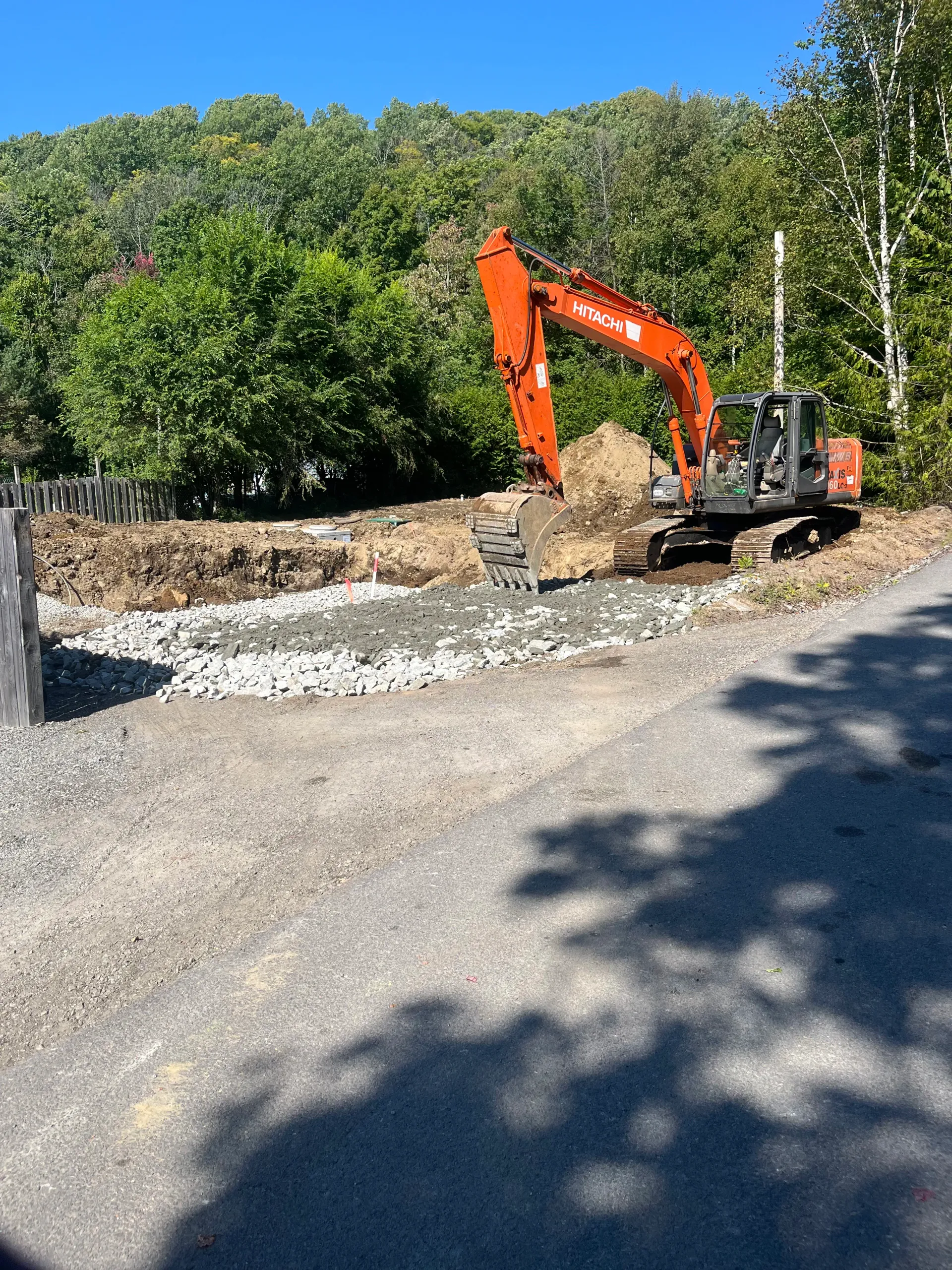 Orange excavator grading gravel at a construction site beside a stone wall and trees