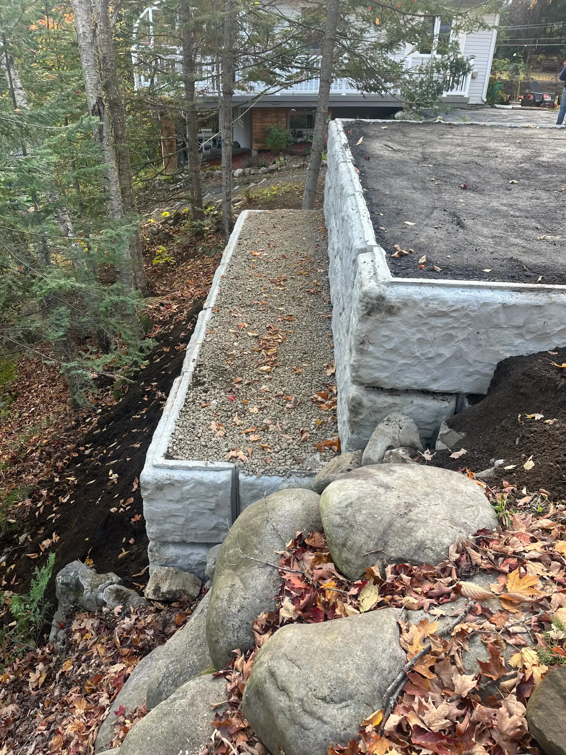 Concrete retaining wall with a narrow gravel path beside rocks and autumn leaves in a wooded yard