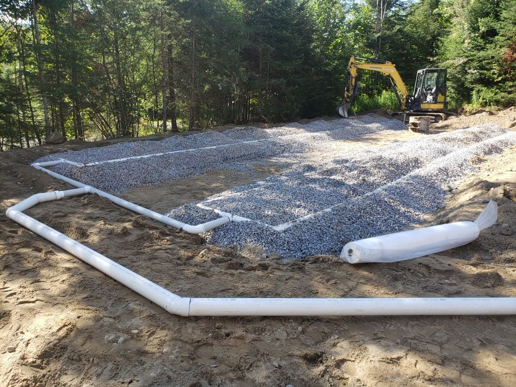 Excavator working on a gravel drainage field beside white PVC pipes in a wooded area