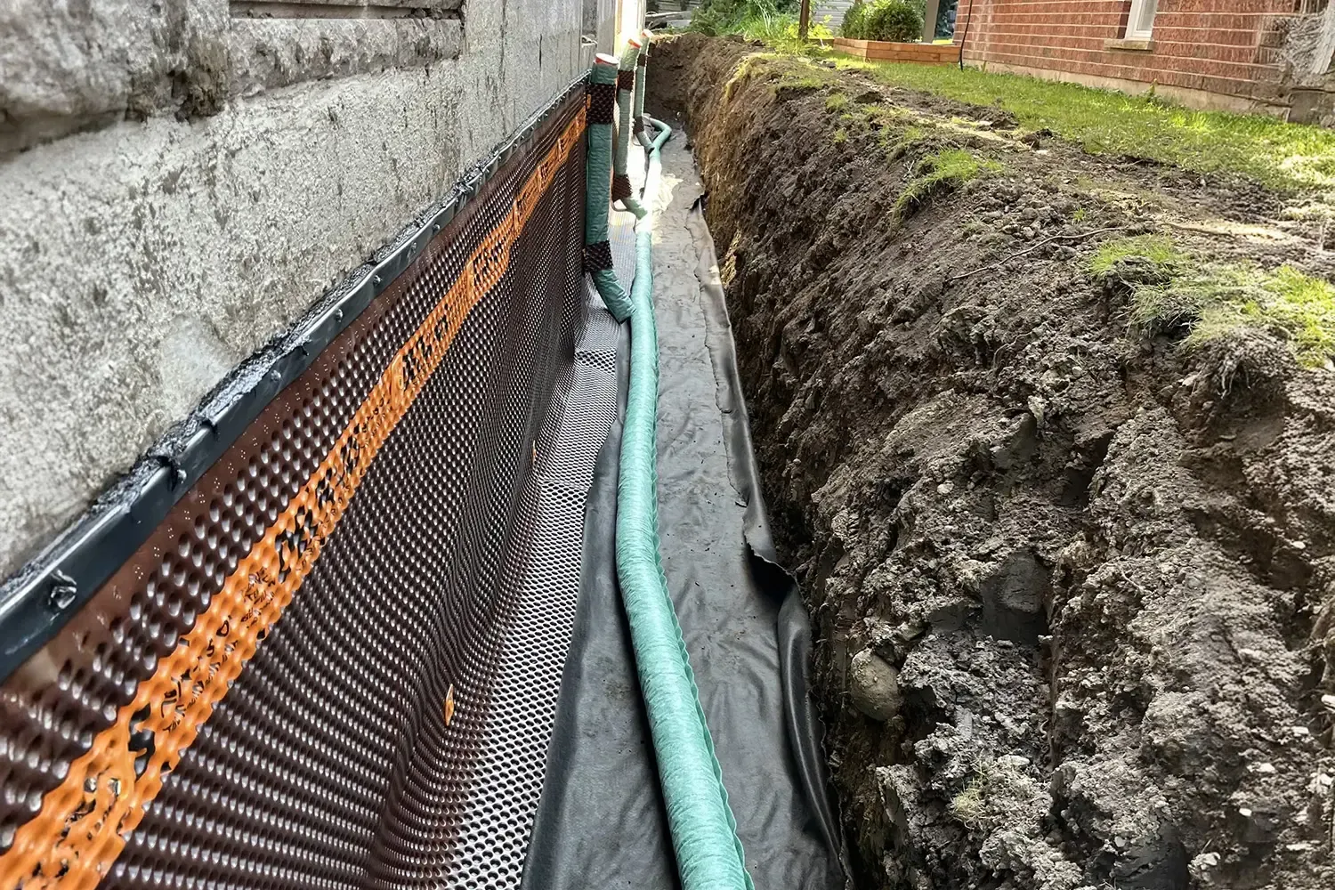 Trenched foundation beside a wall with drainage pipe and black-and-orange waterproofing mesh