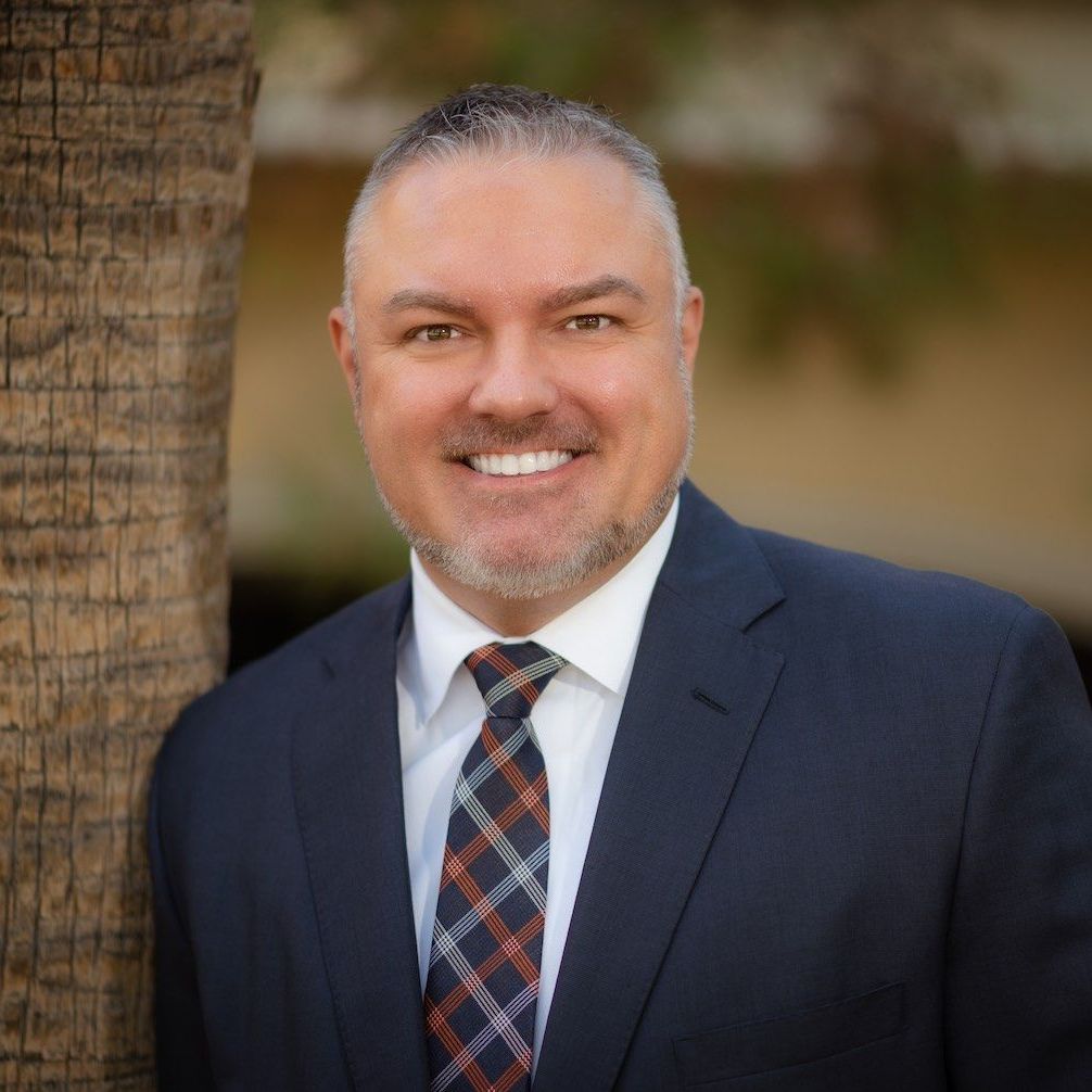 A man in a suit and tie is smiling while leaning against a tree.