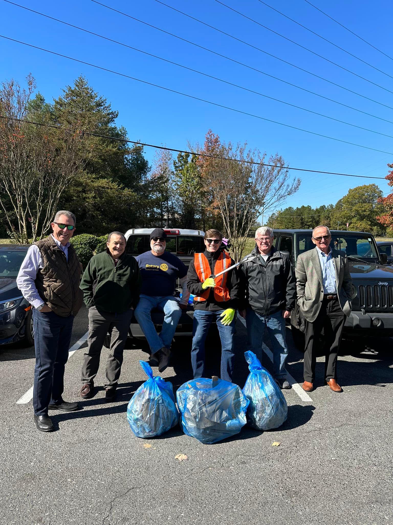 Six people stand by blue trash bags in a parking lot on a sunny day.
