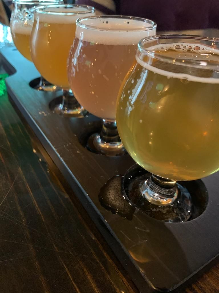 Four small beer tasters in clear glasses lined up on a dark tray at a bar.