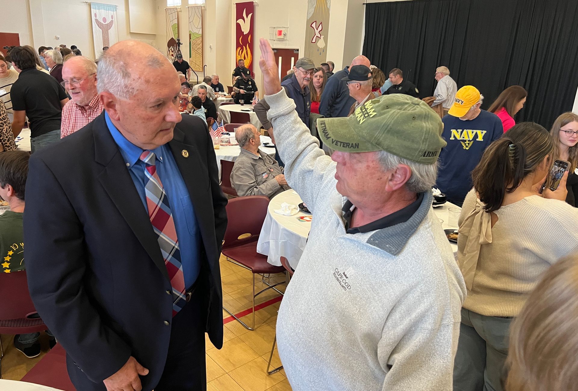Two men talk in a crowded hall; one gestures while wearing a green cap and light sweater.