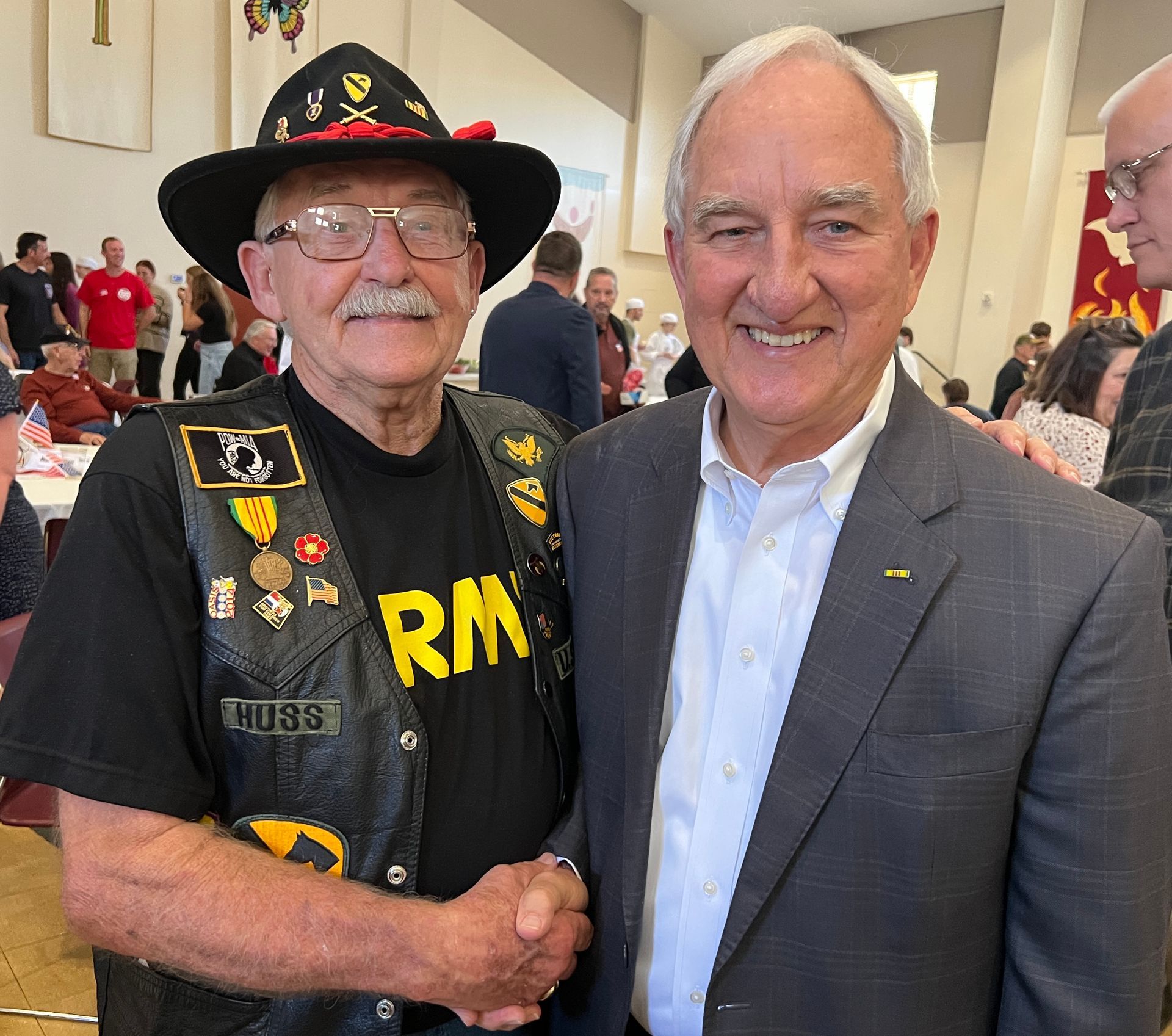 Two men smiling and shaking hands at an indoor gathering, one in a black veteran vest and hat.