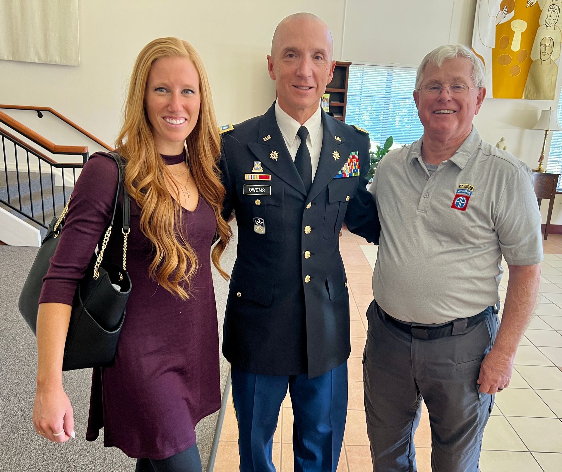 Three people posing indoors, including a uniformed military officer, in a bright lobby with stairs.