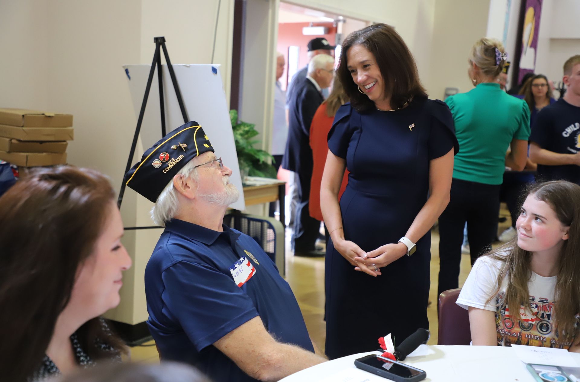 Veteran in cap speaks with seated women at a table during a hallway gathering