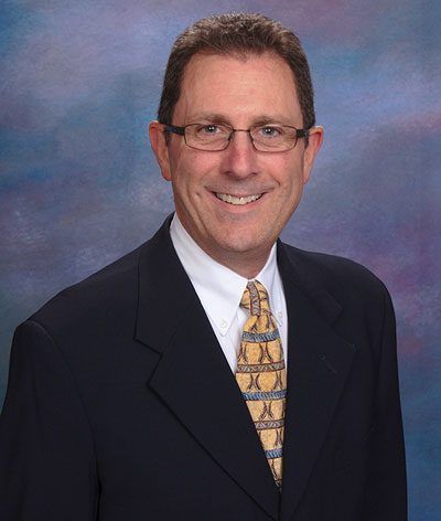 Smiling man in dark suit, glasses, and patterned tie against a blue studio backdrop