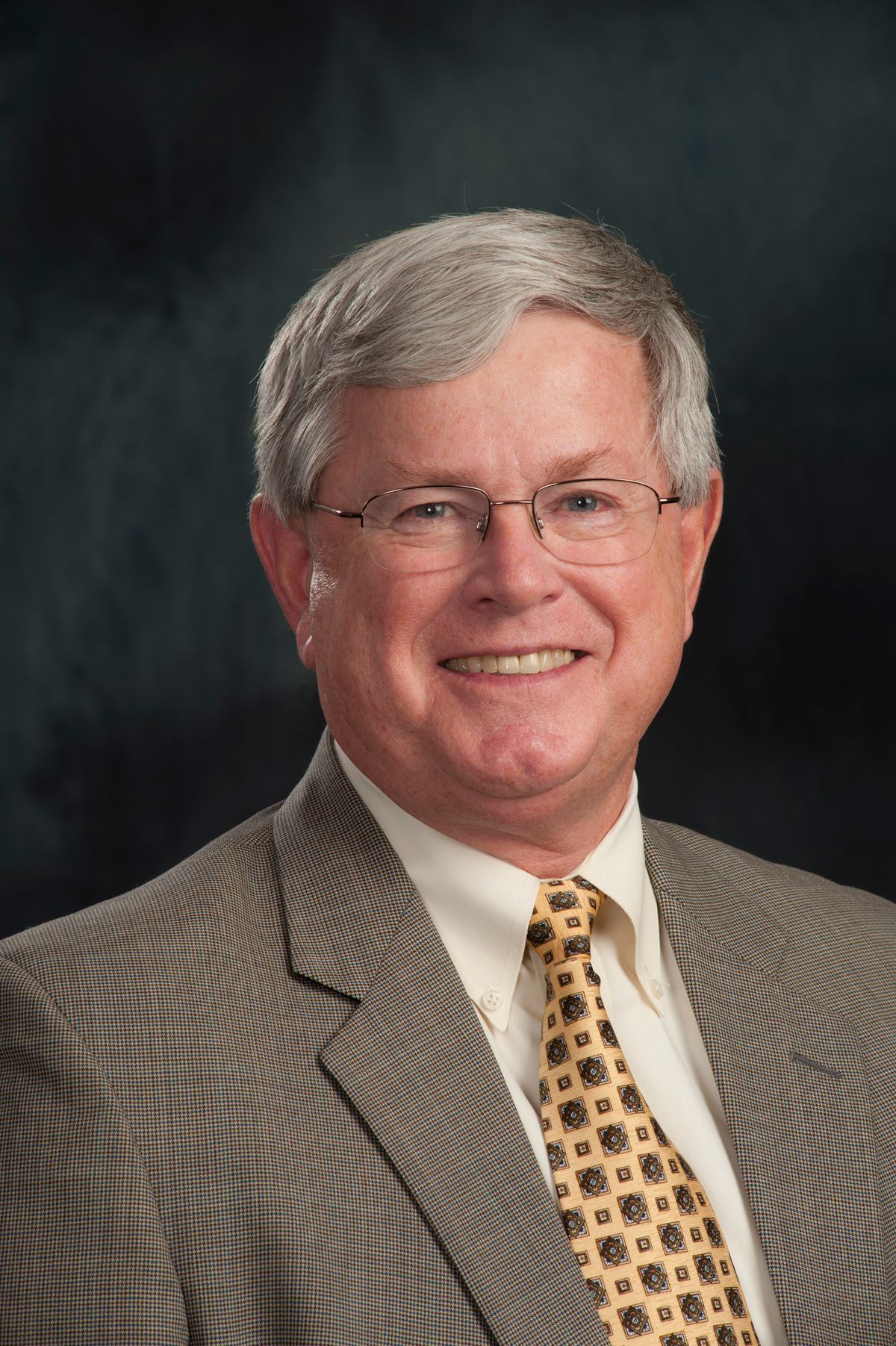 Portrait of a smiling man in a gray suit, white shirt, and patterned tie against a dark studio backdrop