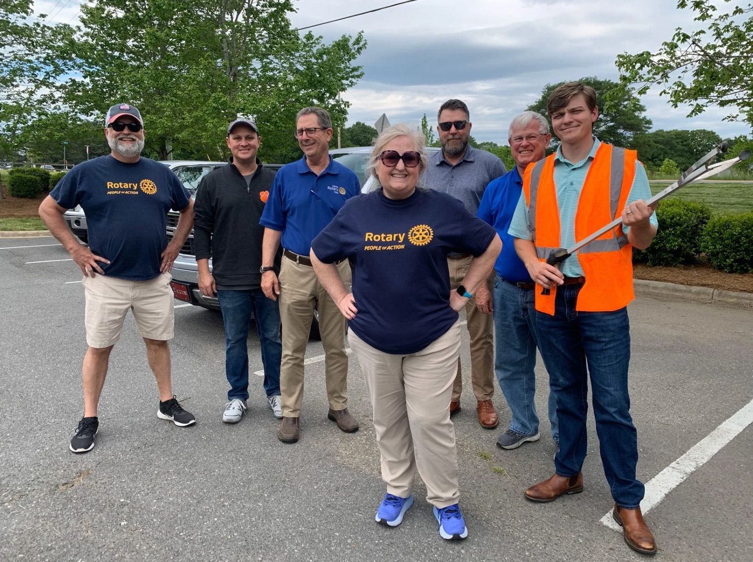Six people posing outdoors on a road, smiling and wearing navy shirts, one in an orange safety vest.