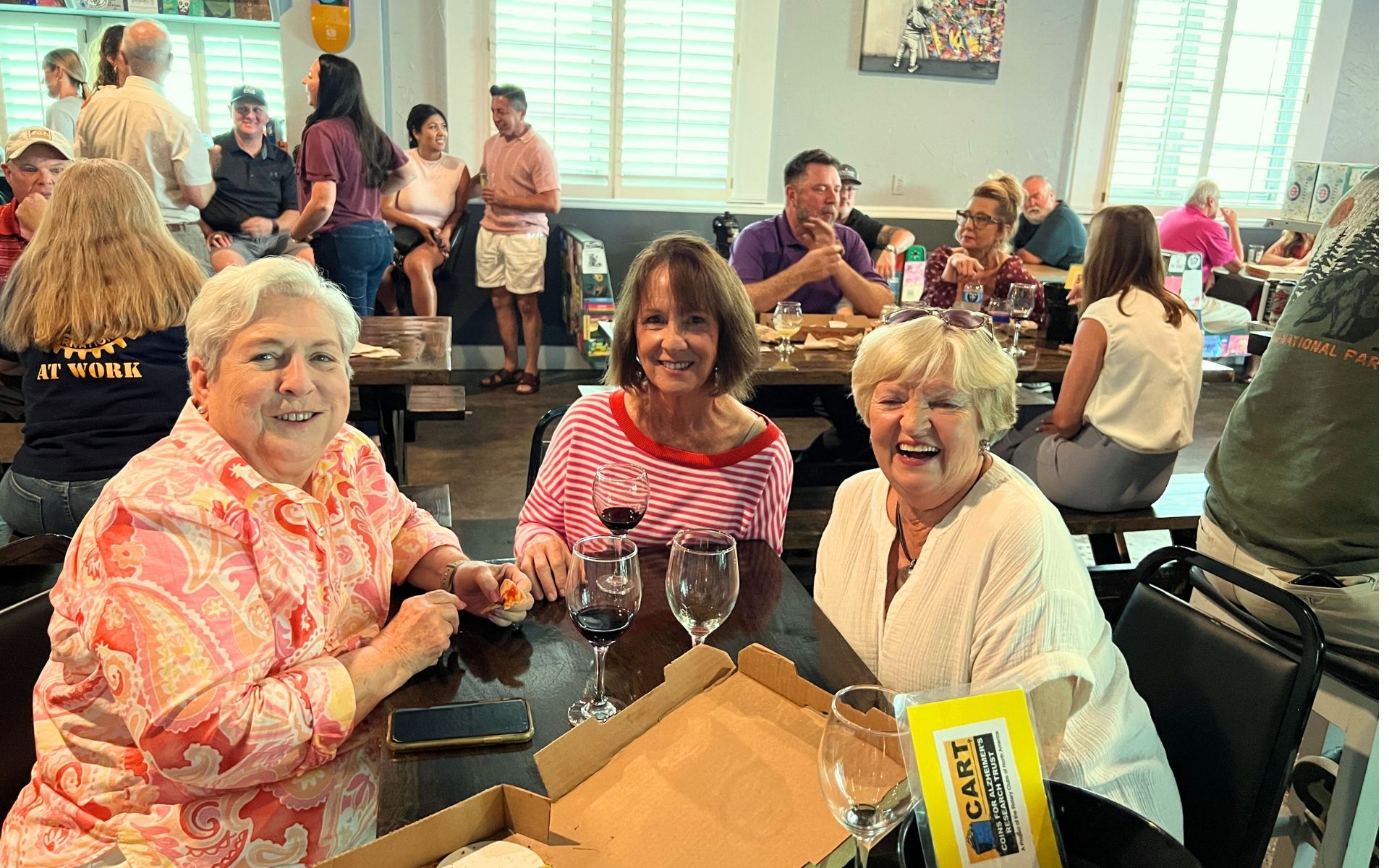 Three people smiling at a table in a bright community room with drinks and snacks