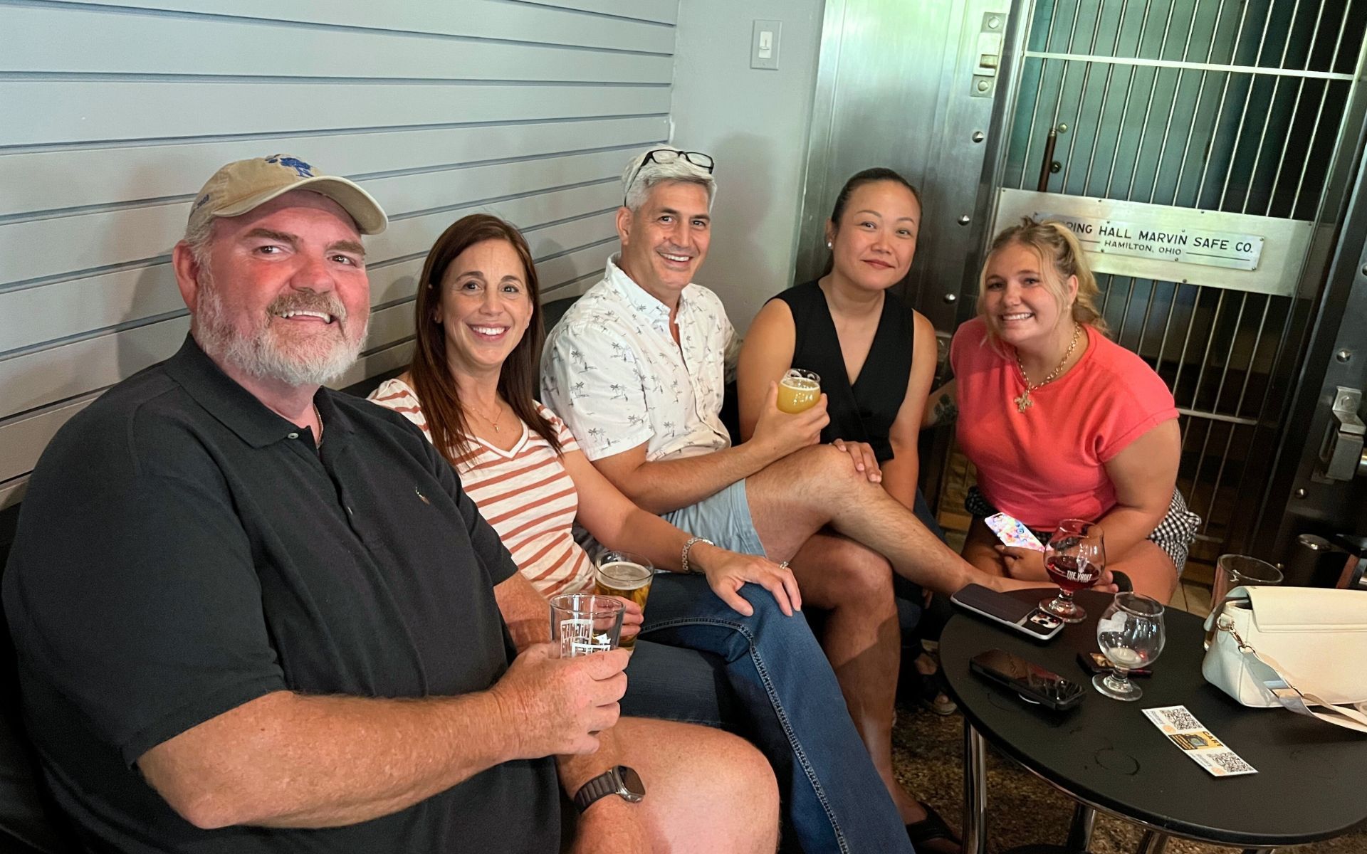 Five people sitting together on a patio, smiling and holding drinks around a small table.
