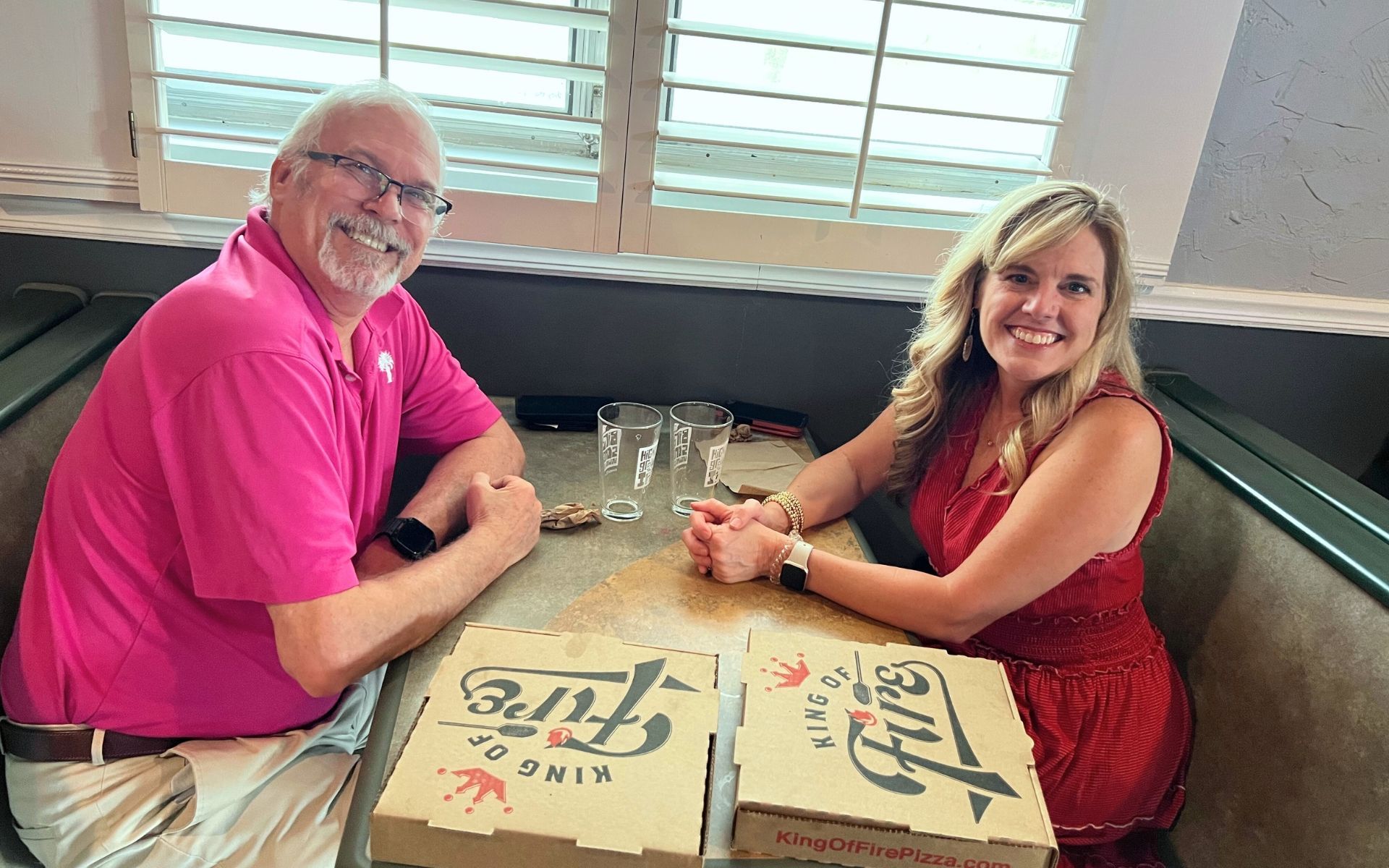 Two people smiling at a restaurant booth with drinks and pizza boxes on the table.