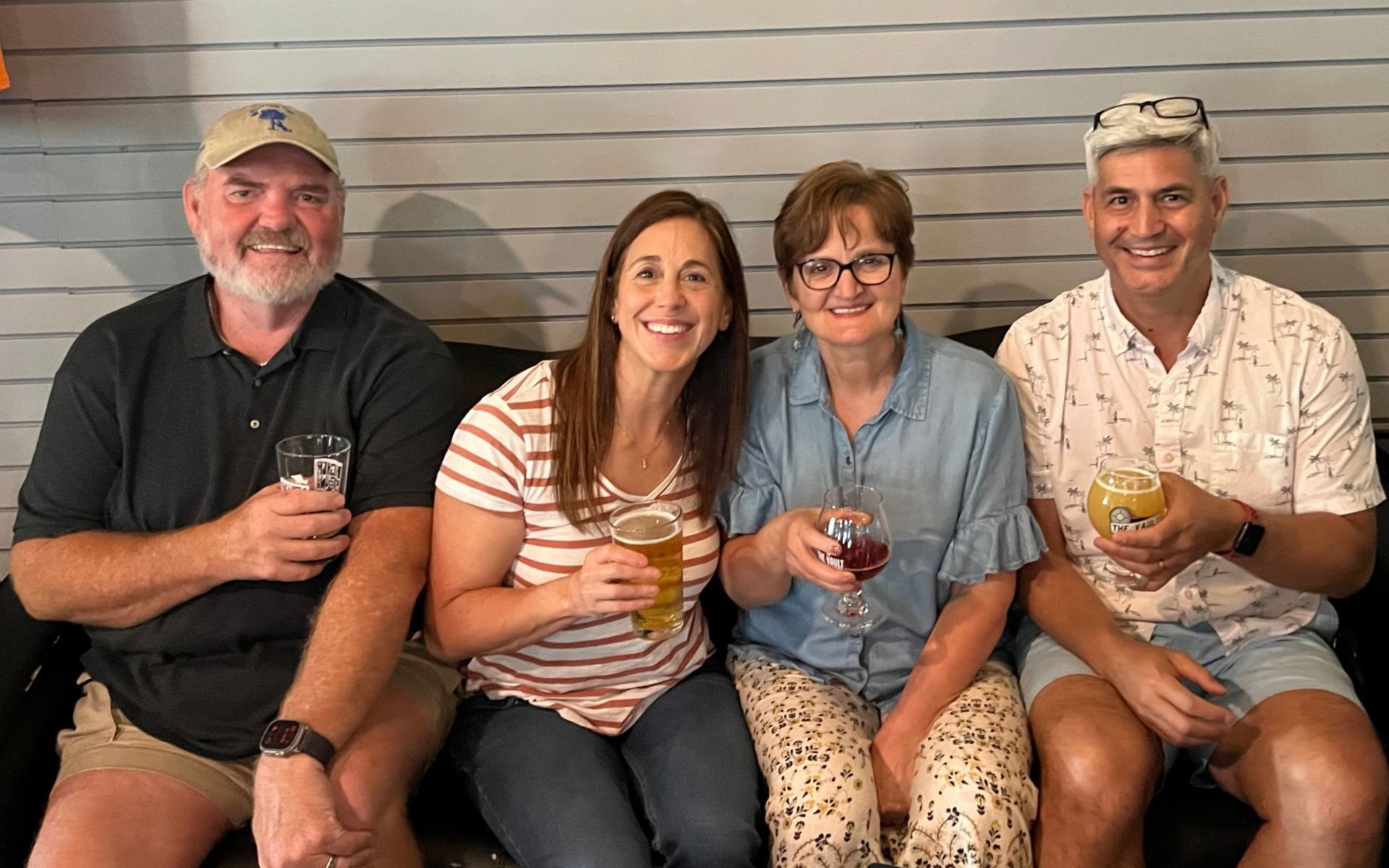 Four friends sit on a bench smiling and holding drinks against a gray siding wall.