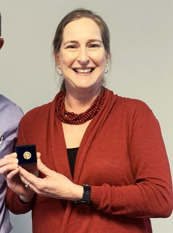Smiling woman in a red cardigan holding a small blue award plaque against a plain wall.