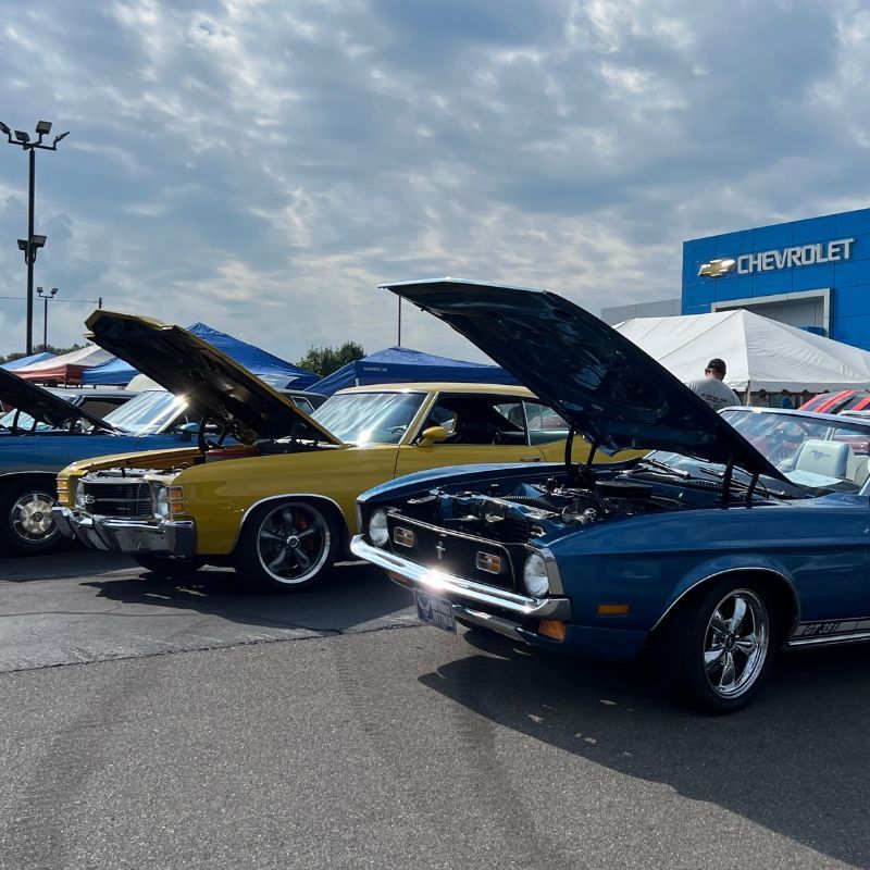 Classic cars with open hoods on display at an outdoor Chevrolet event under a cloudy sky