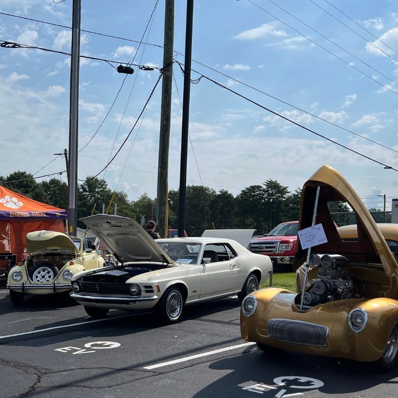 Classic cars on display at an outdoor car show, including a gold hot rod and a white coupe with its hood open.