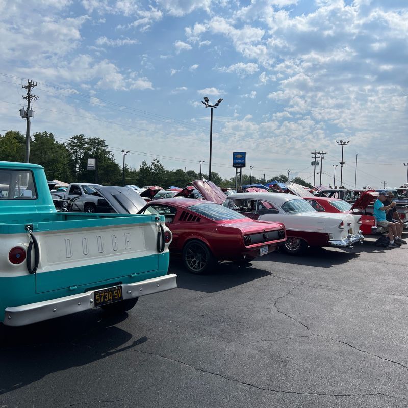 Classic cars parked in a lot, including a turquoise pickup and red sports cars under a cloudy sky