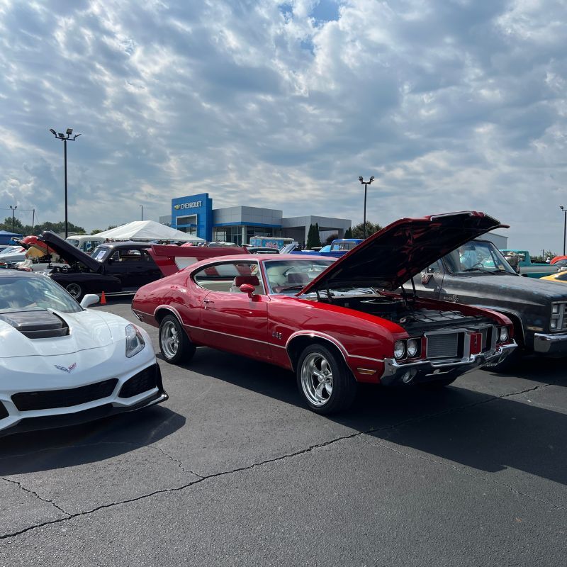 Classic cars on display at an outdoor car show, including a red muscle car with its hood open and a white sports car.