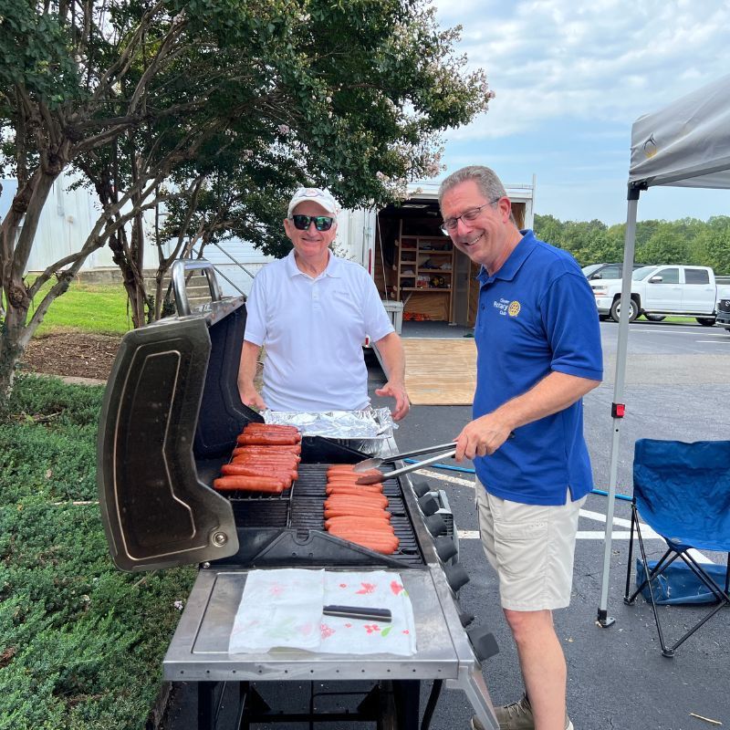 Two men grilling hot dogs outdoors, one smiling in a blue shirt beside a black grill