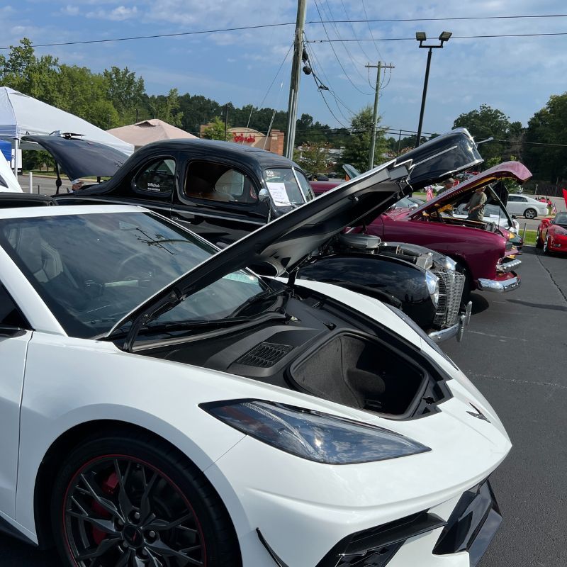 White sports car at an outdoor car show with hood open, other classic cars lined up behind it