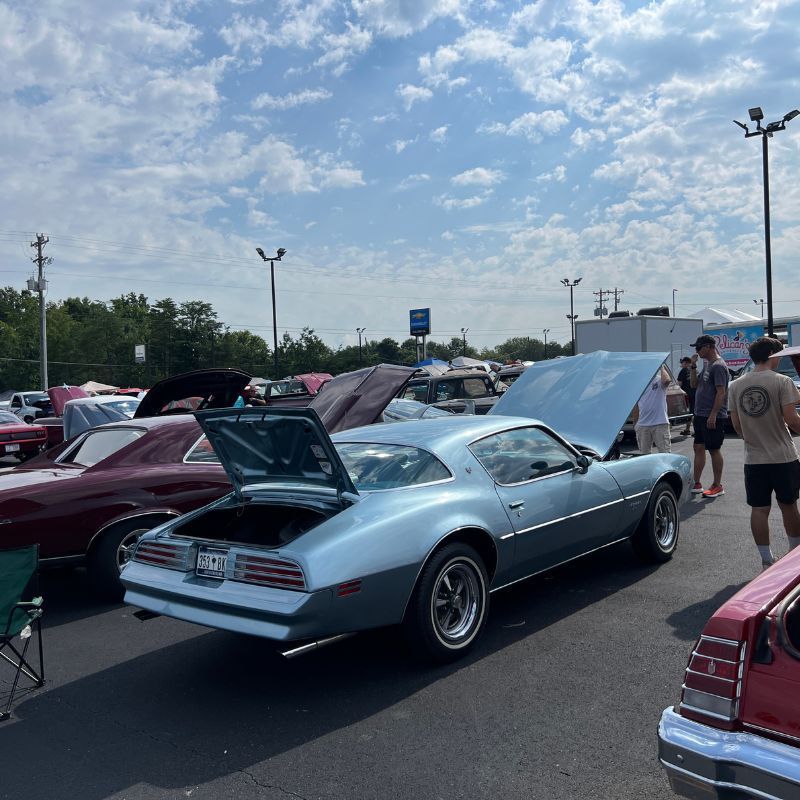 Classic cars on display at an outdoor show, with a light blue coupe in the foreground and open hoods.
