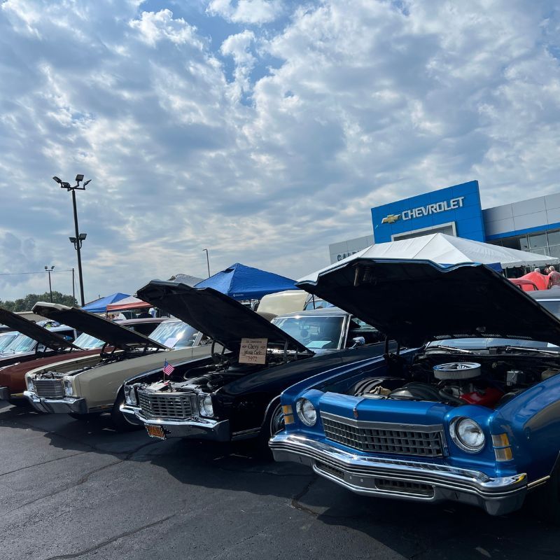 Classic cars on display at a Chevrolet lot, with raised hoods and a blue car in the foreground.