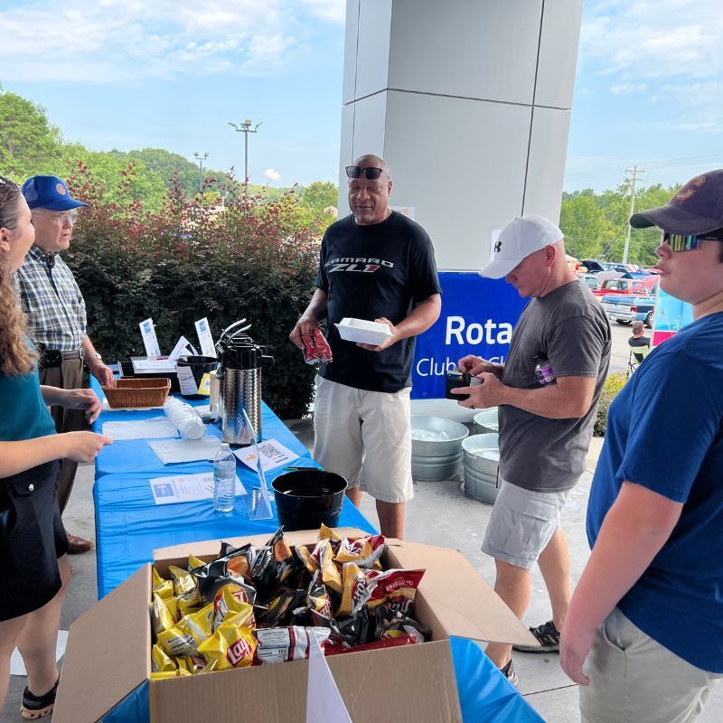 People gathered at a Rotary Club table with snacks and papers outdoors under a covered area.