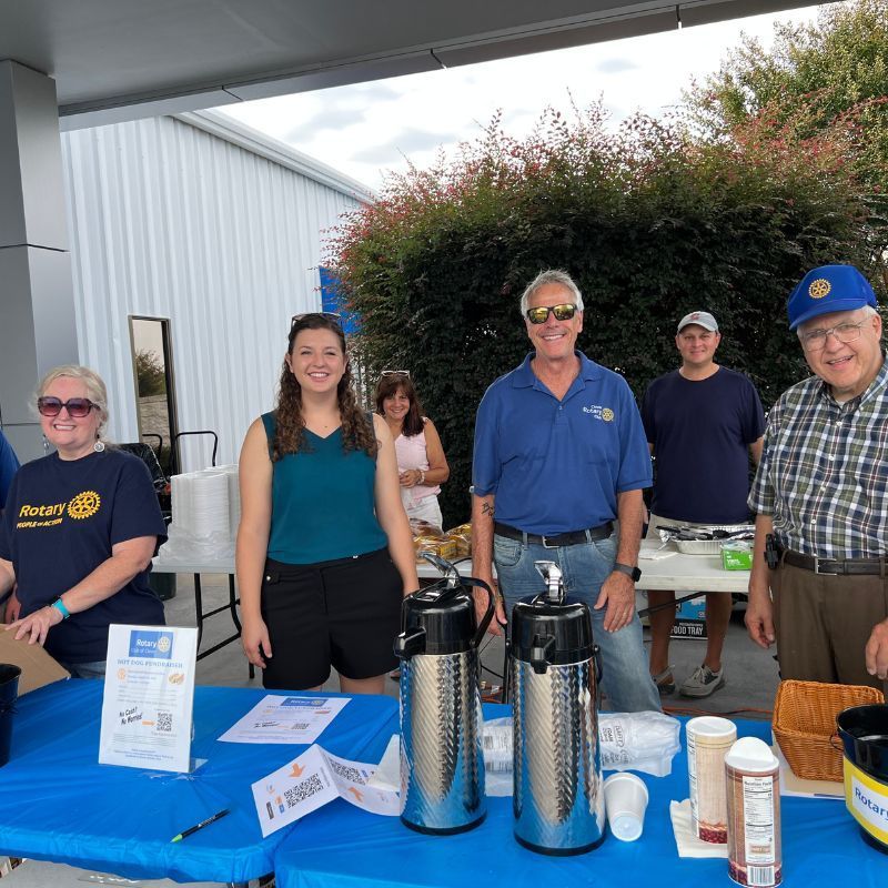 Five people standing behind a blue table with coffee dispensers at an outdoor event under a covered patio