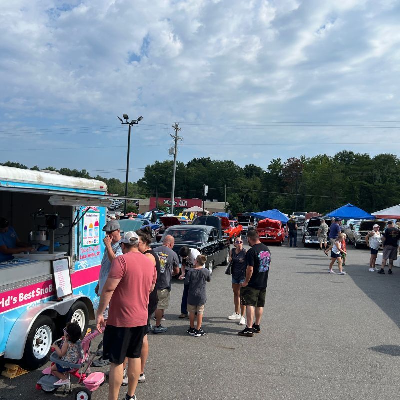 People browsing at an outdoor car show with classic cars, food trucks, and blue tents under a cloudy sky