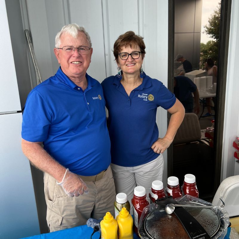 Two people in blue polos stand behind a table with condiments and a grill outdoors.