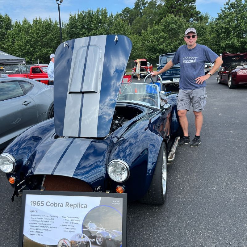Blue 1965 Cobra replica with open hood at a car show; man stands beside it.