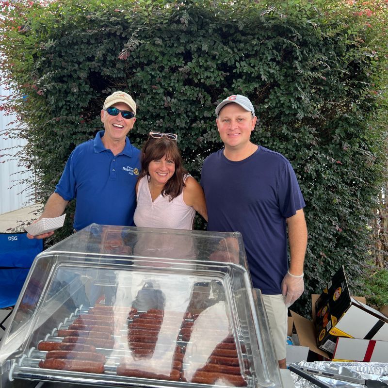 Three people posing behind a display case at an outdoor market, with a hedge in the background.