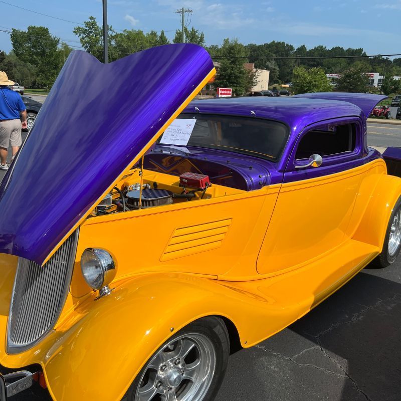 Purple-and-yellow custom hot rod with hood open at an outdoor car show